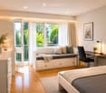 Modern bedroom with a bed, built-in sofa, desk, chair, lamp, and decorative items. Large windows and a balcony offer a view of greenery.
