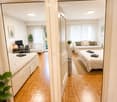 A well-lit, modern furnished room with wooden flooring, a bed, a sofa, a desk with a screen, and a small plant. The room features large windows with white curtains and a view of greenery outside.