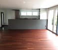 Empty room with wooden flooring, large windows, and a kitchen area visible in the background.