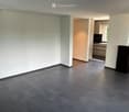 Empty room with dark grey tiled floor and white walls, connected to a modern kitchen area with grey cabinets and a countertop.