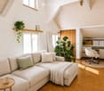 Bright living room with a sectional sofa, coffee table, and a small desk area with a chair. Features large windows, wooden flooring, and decorative plants.