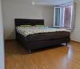 Bright bedroom with wooden flooring, a large bed with a dark headboard and green pillows, and a round ceiling light. The room has large windows with blinds.
