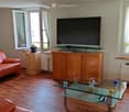 Living room with wooden flooring, large windows, a red leather sofa, a glass coffee table, a TV on a wooden stand, and a small plant on a side table.