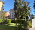 Well-maintained garden with a lawn, trimmed bushes, and tall trees, including a prominent cypress tree. The area is bordered by a paved walkway and a low stone wall, with residential buildings visible in the background.