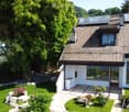 Modern two-story house with solar panels on the roof, large windows, and a well-maintained garden with a stone pathway and various plants.