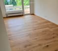 Empty room with light wooden flooring, white walls, large window and sliding glass door leading to a balcony.