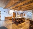 Cozy dining area with wooden beams, exposed wooden ceiling, dining table with chairs, vase with flowers, wooden cabinet, and a small seating area with a sofa and stools.