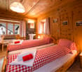 Cozy wooden bedroom with two single beds, red and white checkered bedding, nightstands, a small wooden chair, a mirror, and a sink in the corner.