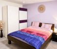 Bedroom with a wooden bed, colorful bedding, nightstand with lamp, vase with dried flowers, and decorative wall art.