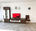 Modern living room with a large TV on a wooden stand, a brown leather chair, a wardrobe, and a glass shelf with decorative items.