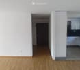Empty hallway with wooden flooring, white walls, and a door leading to another room. A kitchen area is visible at the end of the hallway with white cabinets and a countertop.