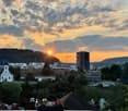 Panoramic cityscape view at sunset, featuring a mix of modern high-rise buildings, historic structures, and lush greenery.