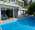Outdoor area with a swimming pool, two striped lounge chairs, and a modern house with a balcony and greenery.