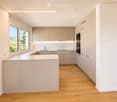 Modern kitchen with light-colored cabinets, a central island, and a sink area. Features wooden flooring, large windows, and built-in appliances.