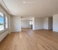 Empty room with wooden flooring, white walls, large windows, and a small cabinet in the background.
