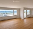Empty room with wooden flooring, large windows offering scenic mountain and lake views, radiators, and a sliding glass door leading to a balcony.