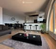 Modern open-plan living and dining area with a grey sofa, black coffee table, dining table with chairs, and a kitchen island with stools. The space features wooden flooring, large windows, and contemporary decor including a plant and flower vase.