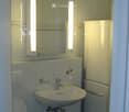 Modern bathroom with white tiles, a sink with a chrome faucet, a toilet, a mirror with built-in lighting, and a white cabinet.