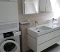 Modern bathroom with a white sink, chrome faucet, large mirror, and a washing machine. The sink has a sleek design with integrated drawers.