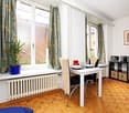 A well-lit dining area with a white dining table, chairs, a potted plant on the windowsill, and a microwave on a small cabinet. The room features wooden flooring and large windows with patterned curtains.