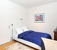 A small, modern bedroom with a white bed, blue duvet, nightstand, chair, and a picture frame on the wall.