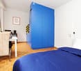 Cozy bedroom with a blue wardrobe, a bed with blue bedding, a small table with a chair, and a framed picture on the wall.