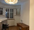 A furnished room with wooden flooring, a large window with blinds, a modern chandelier, a wooden desk, a black chair, and a small table lamp.