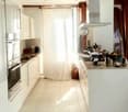 Modern kitchen with white cabinets, stainless steel oven, countertop, and various appliances including a blender. Light-colored tiled floor and large window with white curtain.
