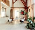 Open-plan living and dining area with exposed wooden beams, white tiled floor, modern furniture including a dining table, chairs, and a kitchen counter. Features large windows, pendant lighting, and decorative elements like a surfboard and potted plants.