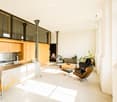 Open-plan kitchen and living area with modern design, featuring a kitchen island, stove, cabinets, a chair, a coffee table, a stool, plants, and a sofa.