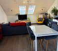 Modern living room with wooden flooring, a grey sofa, a white dining table with four chairs, a yellow armchair, a television, and several plants.