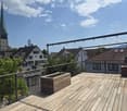 Wooden balcony with metal railing, overlooking a town with a church tower and surrounding houses.