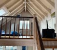 Loft-style room with exposed wooden beams and slanted ceilings. Features a raised sleeping platform with a sofa and table, a kitchen area with a counter and sink, and a TV on a stand.