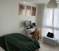 Modern bedroom with a bed covered in green duvet, a desk with a laptop and monitor, a chair, and a small white bench with a black bag on it. The room features light wooden flooring and white walls with framed artwork.