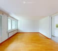 Empty room with wooden flooring, white walls, large windows, and a small kitchen area visible in the background.