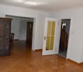 Bright hallway with wooden flooring, white walls, and open doors leading to other rooms. Features a decorative cabinet, a chair, and a staircase visible in the background.