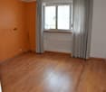 Empty room with wooden flooring, orange accent wall, large window with curtains, radiator, and a broom leaning against the wall.