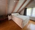 Cozy attic bedroom with wooden slanted ceilings, a double bed with patterned bedding, a nightstand with a lamp, a wooden chair, and a window with dark curtains.