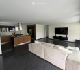 Modern open-plan living and dining area with a large sectional sofa, flat-screen TV, sleek kitchen island with wooden cabinets, and a flower vase on the counter.