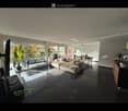 Modern living room with large windows, dark tiled floor, beige sectional sofa, coffee table, dining area with chairs and table, television, and decorative plants.
