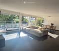 Modern living room with large windows, a sectional sofa, a coffee table, a dining area with chairs, and pendant lights.