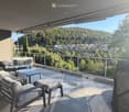 Balcony with modern furniture, including chairs, a table, an ottoman, and a barbecue, overlooking a scenic valley and town.