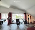 Bright living room with wooden flooring, large windows, and a balcony. Features a sofa, chairs, a table, a fireplace, and decorative statues.