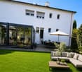 Well-maintained garden with a green lawn, a modern outdoor seating area featuring a sofa, coffee table, and umbrella, along with a dining set under a large umbrella near the house.