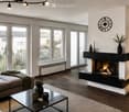 Modern living room with a sectional sofa, coffee table, fireplace, wall-mounted TV, floor lamp, and decorative vases. Large windows and a balcony offer a view of the outside.