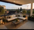 Modern balcony with a white sectional sofa, coffee table, candles, and potted plants, overlooking a cityscape with trees and buildings.