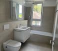 Modern bathroom with a white sink, toilet, bathtub, and a large window providing natural light. The walls are tiled in a neutral color, and there is a mirror above the sink.