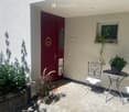 A small outdoor seating area with a metal chair, round table, and potted plants, adjacent to a red front door and window with a small plant on the sill.