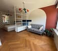 A modern, well-lit room with a grey sofa, a bed, shelves, and a potted plant near the window. The room features wooden flooring and colorful walls.
