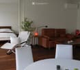 A modern, open-concept living and bedroom space with wooden flooring, a large bed, a brown leather sofa, a white folding chair, a small side table with a lamp, and a potted plant.